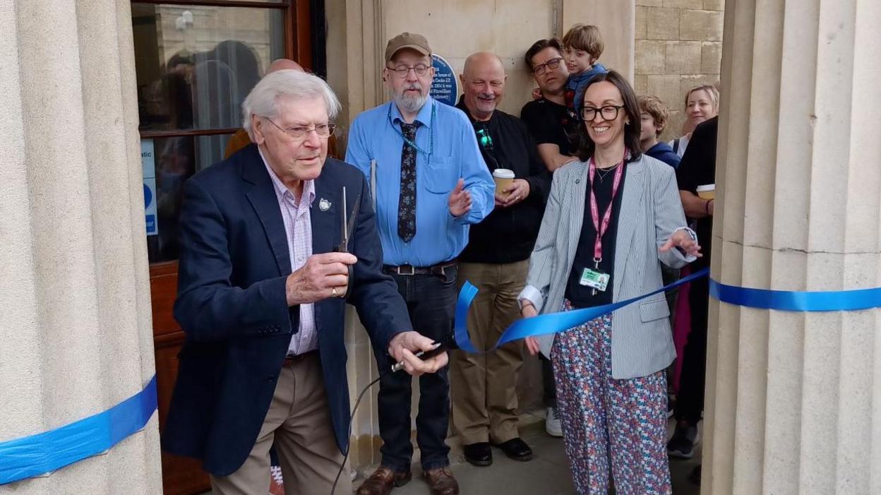 A white-haired man in his 80s, wearing a dark blue jacket, a light blue shirt and light blue trousers, holds up a pair of scissors having just cut through a ceremonial dark blue ribbon tied between two columns at the ornate entranceway into a public building. Various other people stand around watching and applauding.