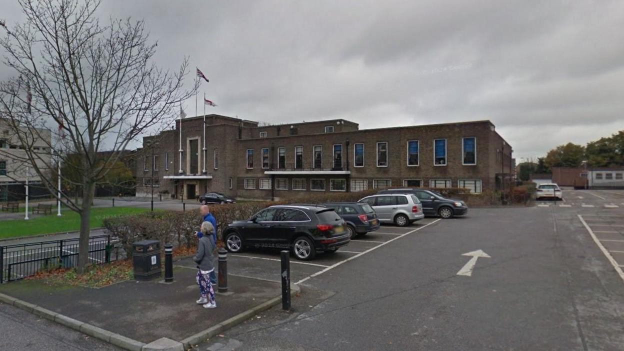 Havering Town Hall, a low-rise brick building, as seen from the car park with two pedestrians