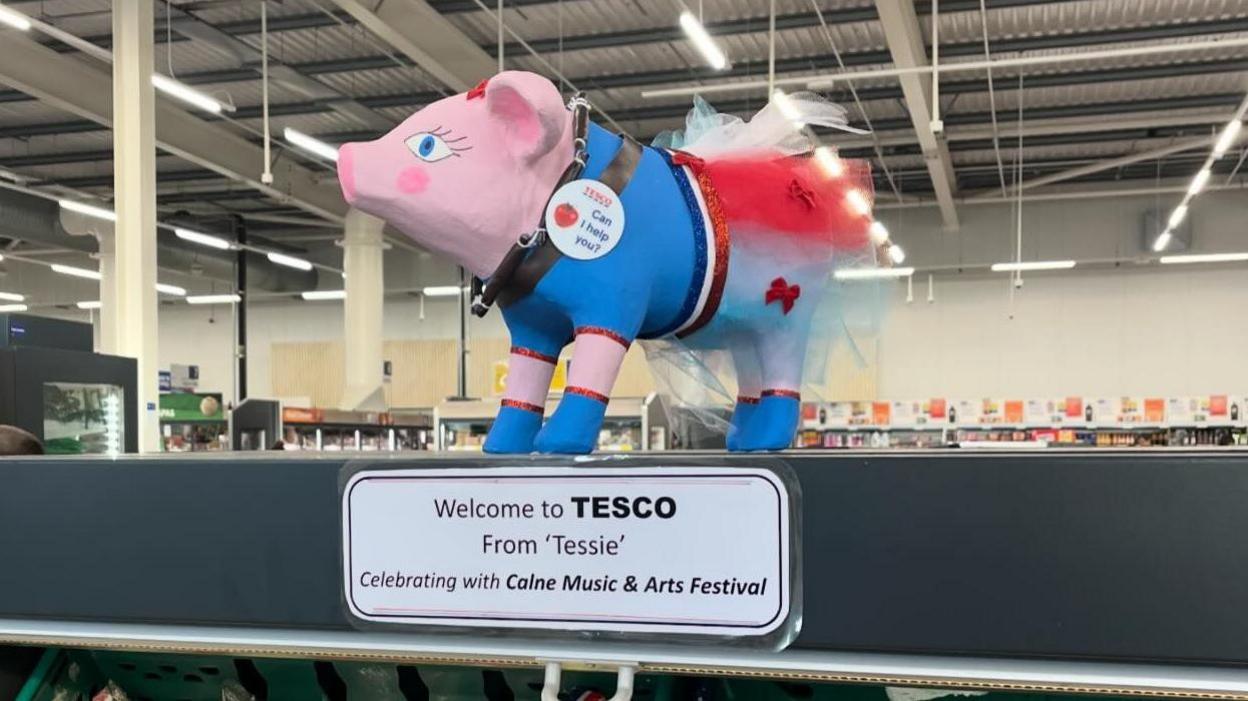 A cardboard pig sculpture, painted pink mostly but also with the Tesco colours of red and blue. This pig has also been given a tutu and little blue feet. A note underneath reads: Welcome to Tesco from Tessie, celebrating with Calne Music & Arts Festival.