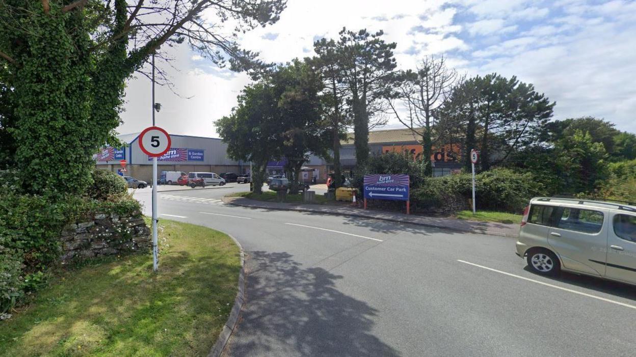 Google Street View image of the entrance to the B&M car park in Penzance. A sign to the car park and entrance with blue and orange B&M branding can be seen along with a 5-miles per hour speed limit sign. B&M and Halfords stores are in the background and are partially blocked from view by trees. A silver car is on the right of the image at the give way point.