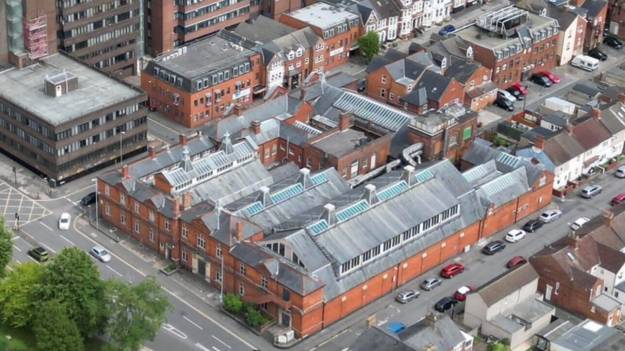 Aerial image of a large building with red brick sides and grey top. Parked cars and a road outside can be seen.
