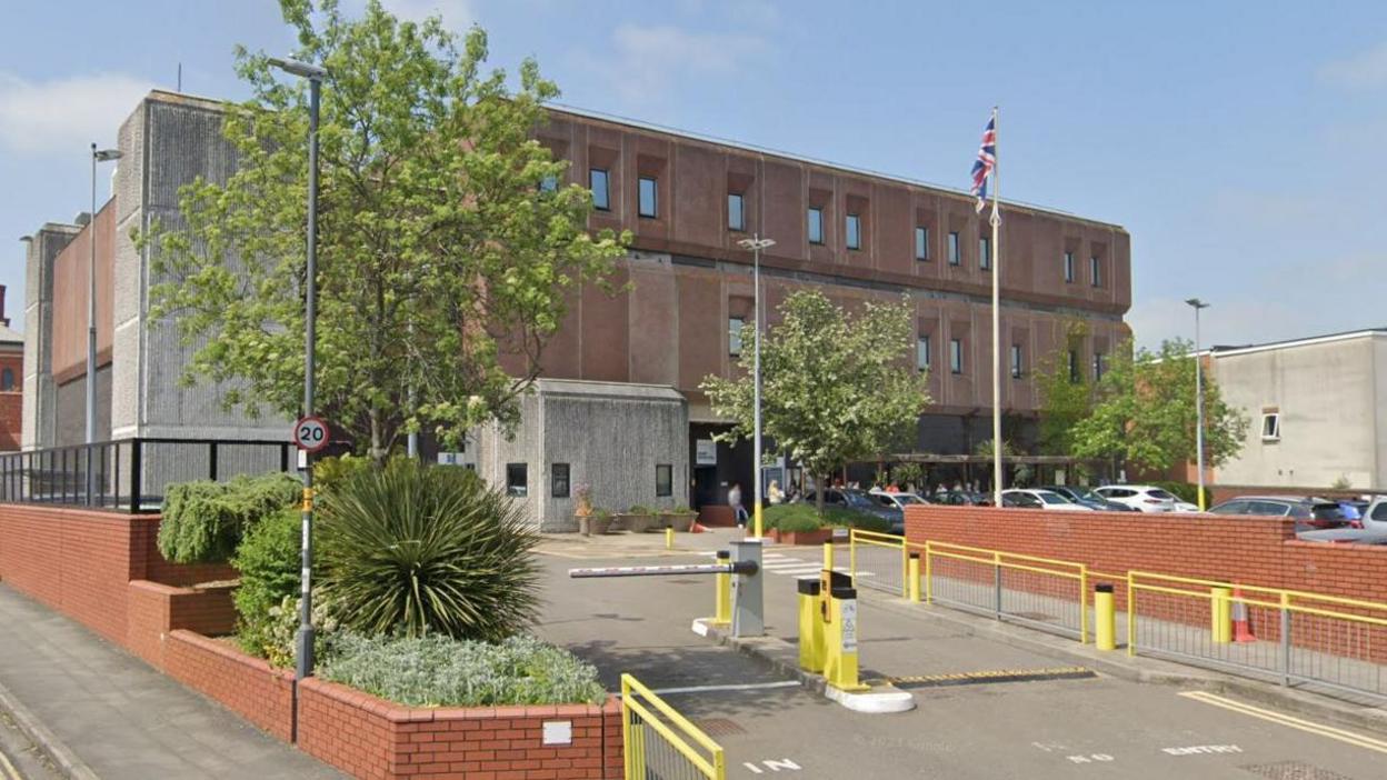 A Google street view screenshot showing the entrance into HMP Bristol prison. It is a large boxy looking building with inset windows. There are yellow railings and an electric barrier leading into the car park. There is a red brick wall that surrounds the perimeter of the prison along with various trees and bushes.