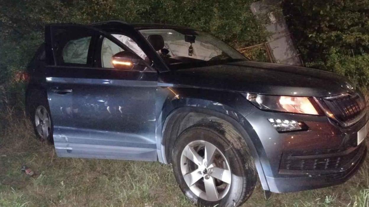 A car resting on grass at the side of the road with the lights still on and clear damage to its doors