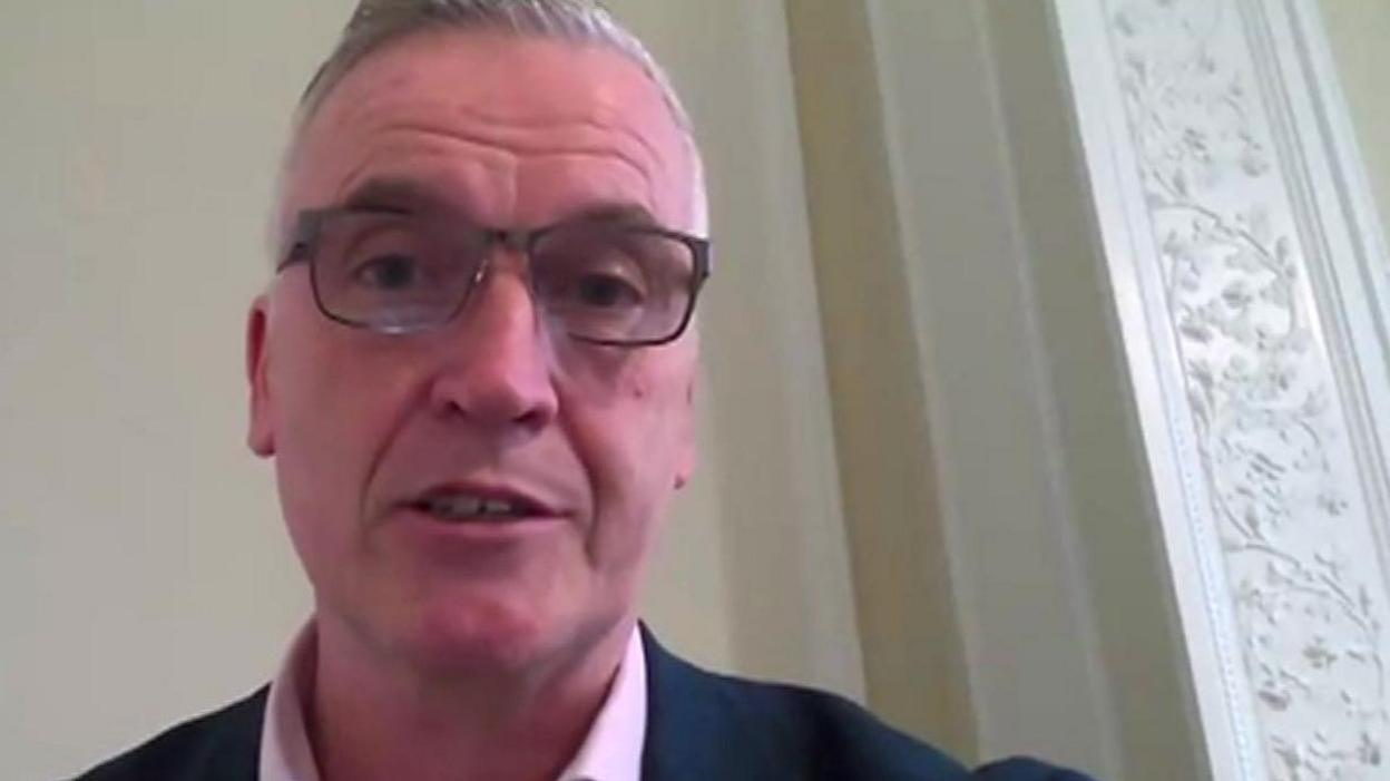 Zoom screengrab of Gareth Roberts in a council room. He wears a pink shirt, dark blue suit and glasses. His face is quite pink.