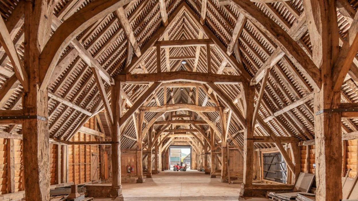 Interior view of a vast vaulted barn made entirely of wood. The structure resembles the inside of a church, with supporting pillars either side of a central aisle.