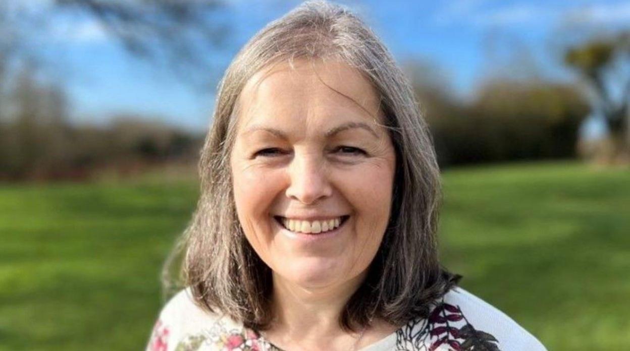 A woman with grey hair and wearing a white top including partially red and green flower patterns is smiling at the camera. A field behind her is blurred in the background.