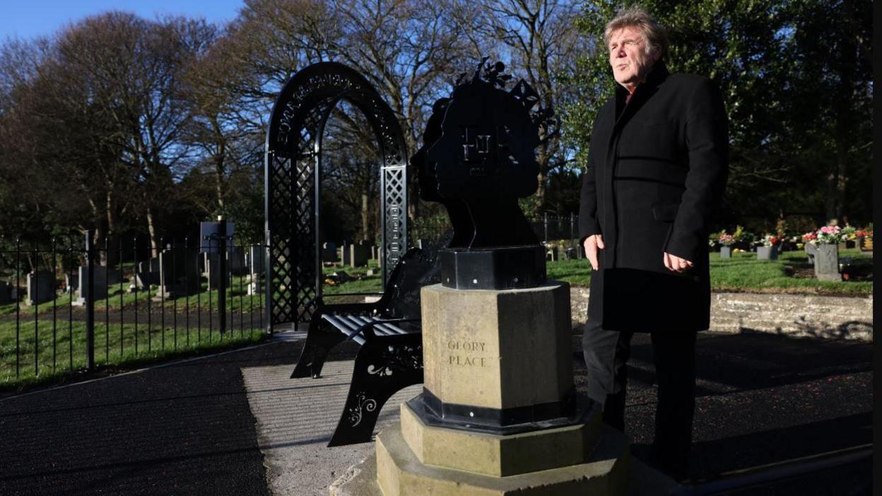 Councillor Malcolm Robinson, a middle aged man with a serious expression and wearing a dark overcoat, standing next to metal bust of the late Queen Elizabeth II on a stone plinth. Just to the left are metal railings and a wrought metal arch, with gravestones seen behind.