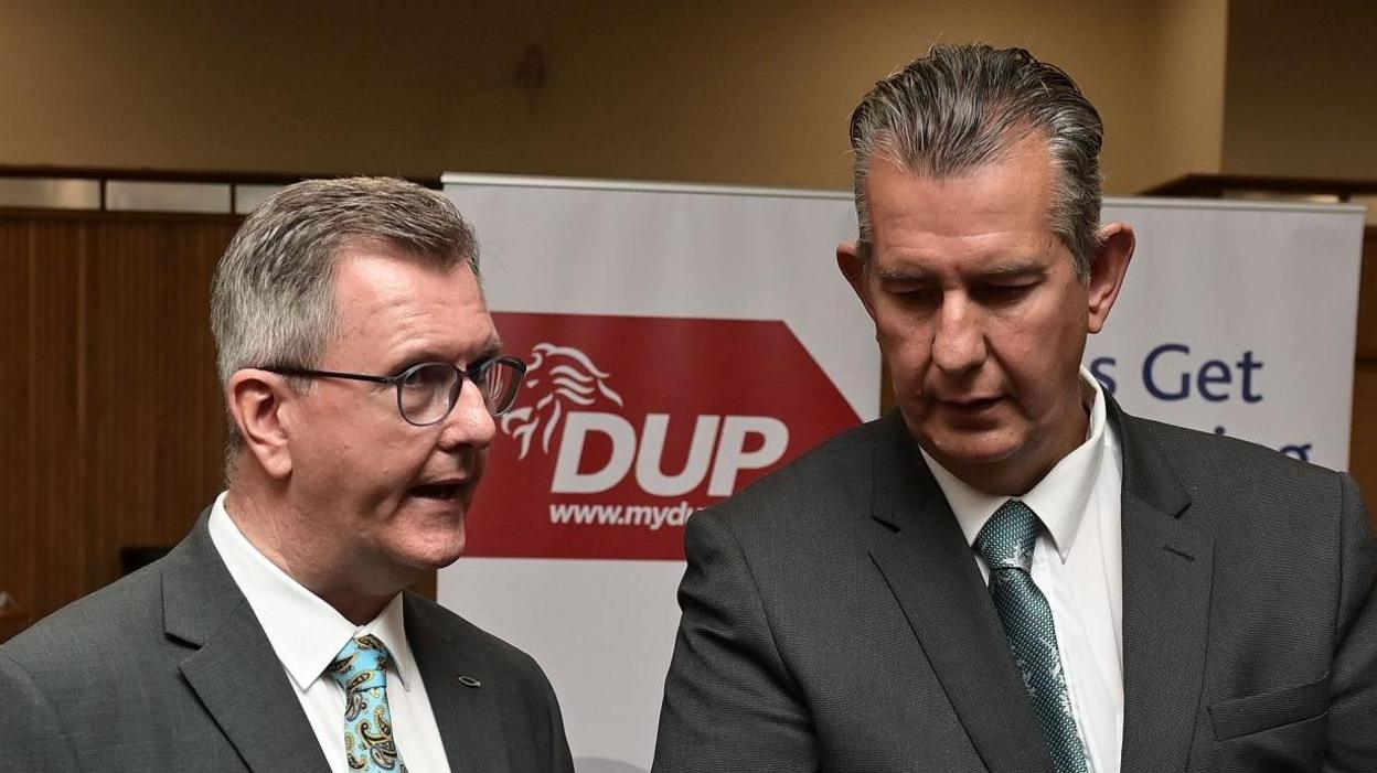 Jeffrey Donaldson and Edwin Poots pictured side by side. Both wearing grey suits. DUP poster in background.
