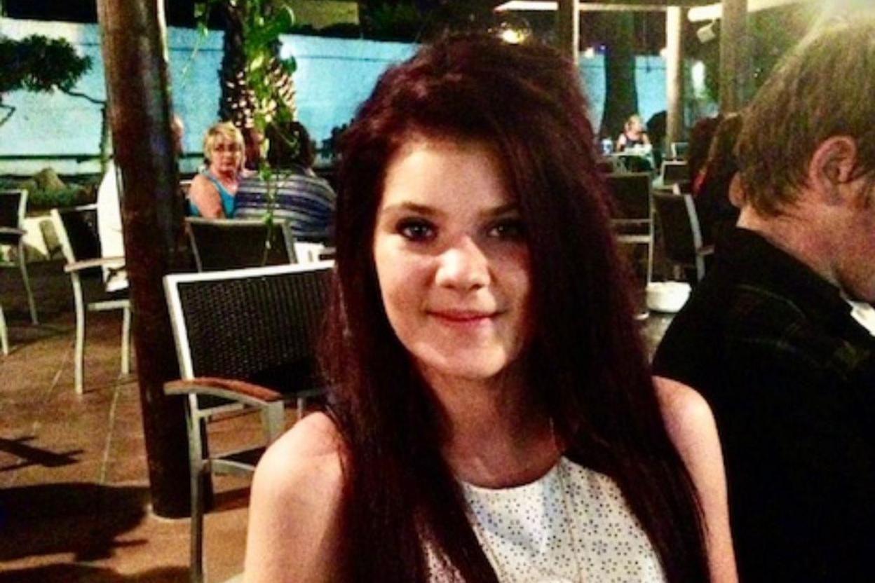 An upper body view of a young woman with long brown hair in a white sleeveless dress sitting in an outdoor dining area and smiling at the camera.