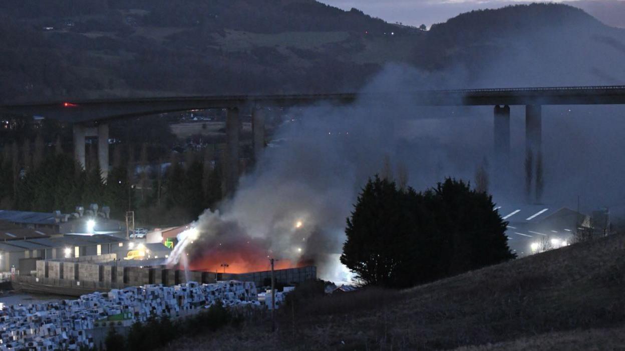 A large fire in a building producing smoke which is drifting upwards to a bridge