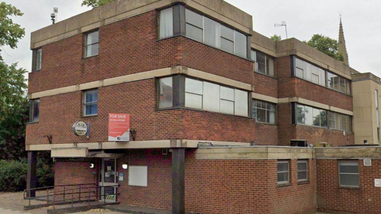 Kettering's former police station on London Road. It is a brutalist-style, three-storey red brick building, with large windows on the top floor. It has a "For Sale" board above the front door.