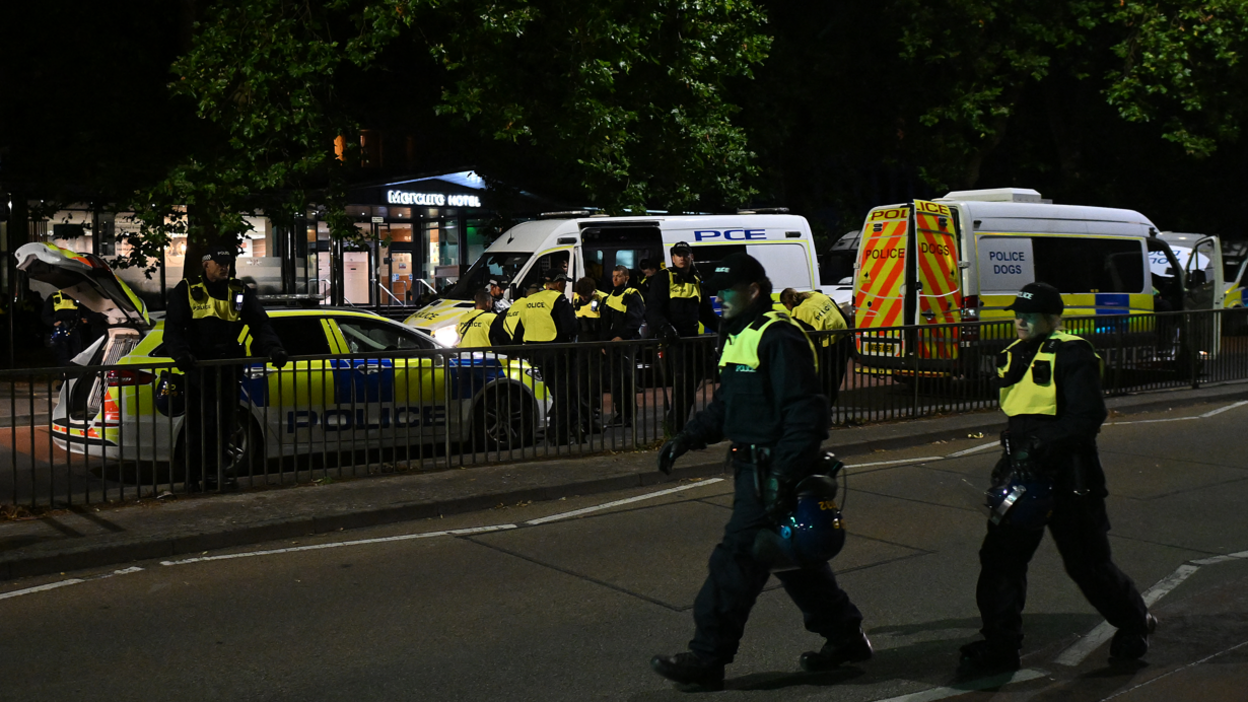 Two police officers walking across the road, with more in the background standing beside a police car and two vans. 