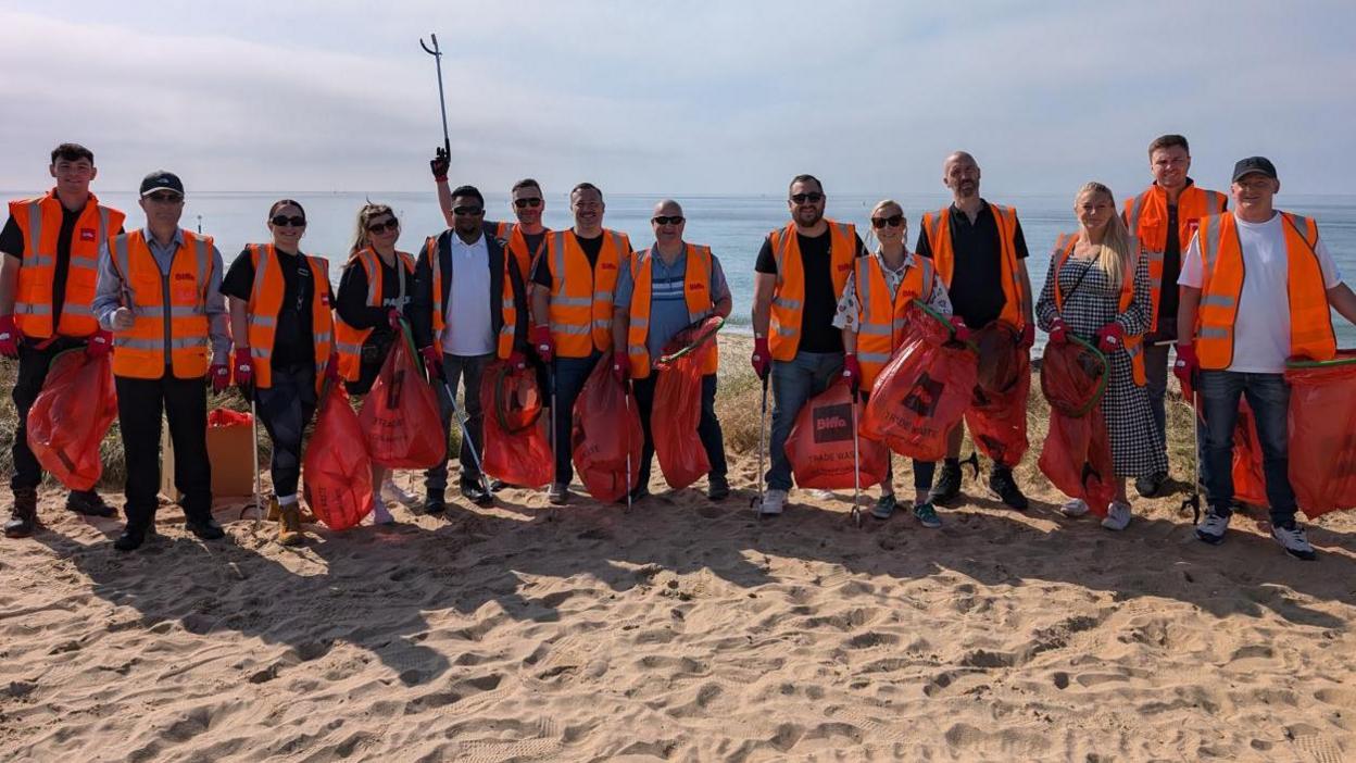 Fourteen men and women volunteers in orange hi viz jackets at a beach on a sunny day. They are smiling at the camera. Everyone is holding a collection bag and some are holding litter pickers. The sea could be seen behind them.