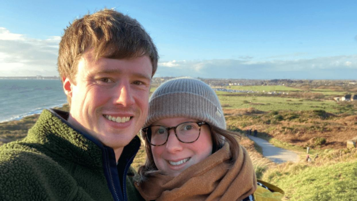 A man and a woman looking at the camera, with a coastline and countryside in the background. The man is wearing a green fleece and is smiling, while the woman next to him is wearing a brown scarf, grey hat and glasses. She is also smiling.