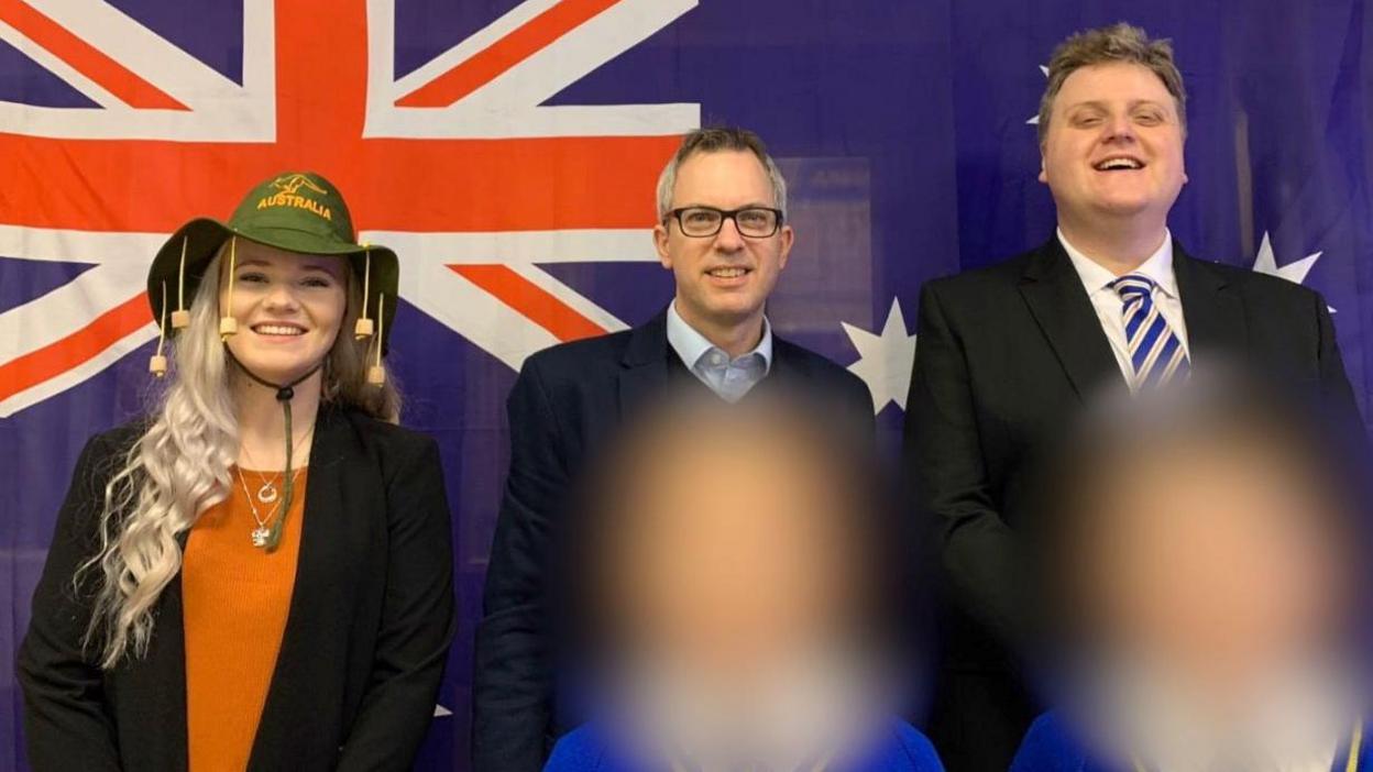 Hannah Gidman stood in a line next to a man and Gregory Hill on a stage. Hannah is wearing a green hat with corks hanging off it and branded with the word "Australia". The two men are wearing suits. They are all stood before a Union Jack flag. 