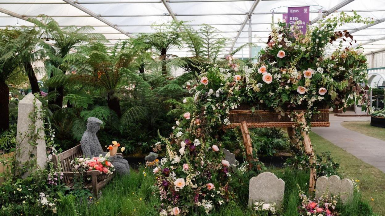 A flower display with a sculpture of a man sat on a bench with a dog at his feat and an elevated willow coffin with flowers bursting out of it. Long grass and a graveyard can be seen below the coffin.