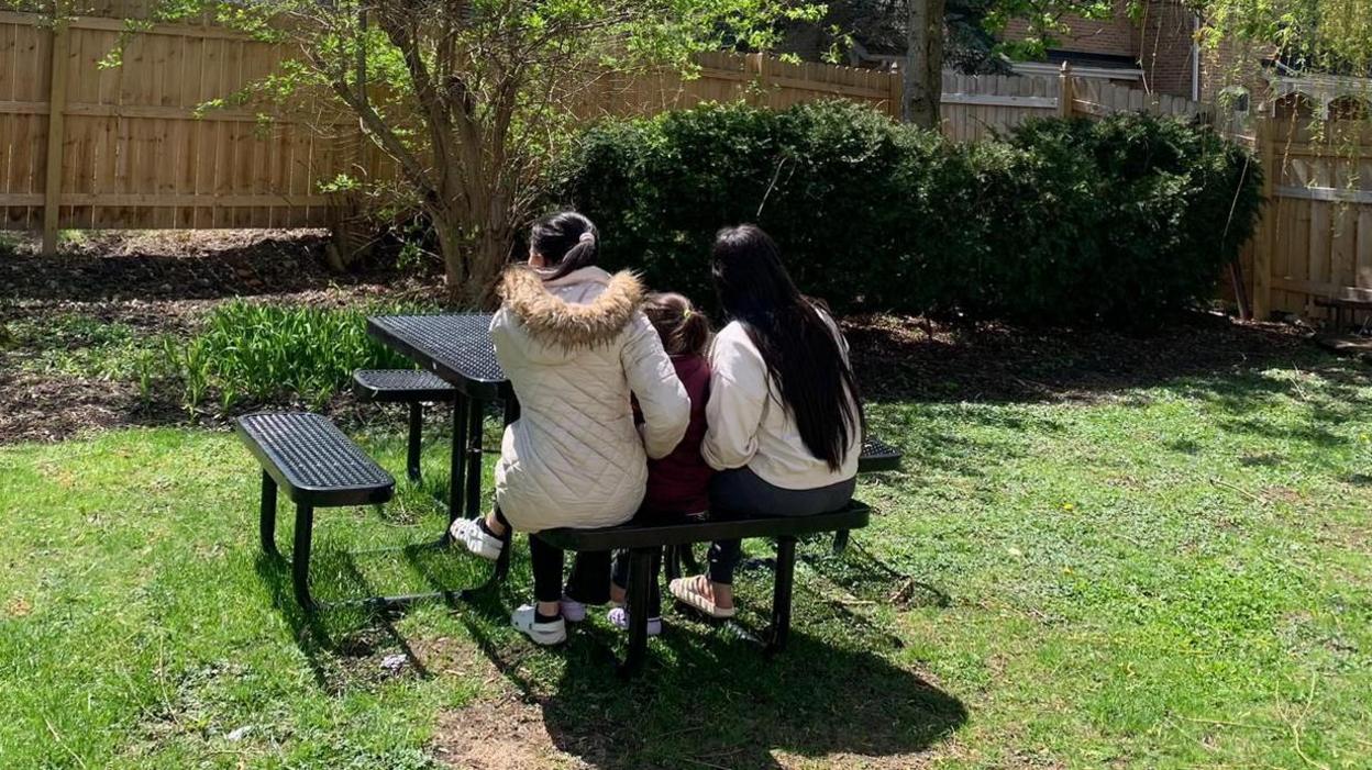 Aracely and her two daughters are sitting at a picnic table in a garden, but we only see them from their back