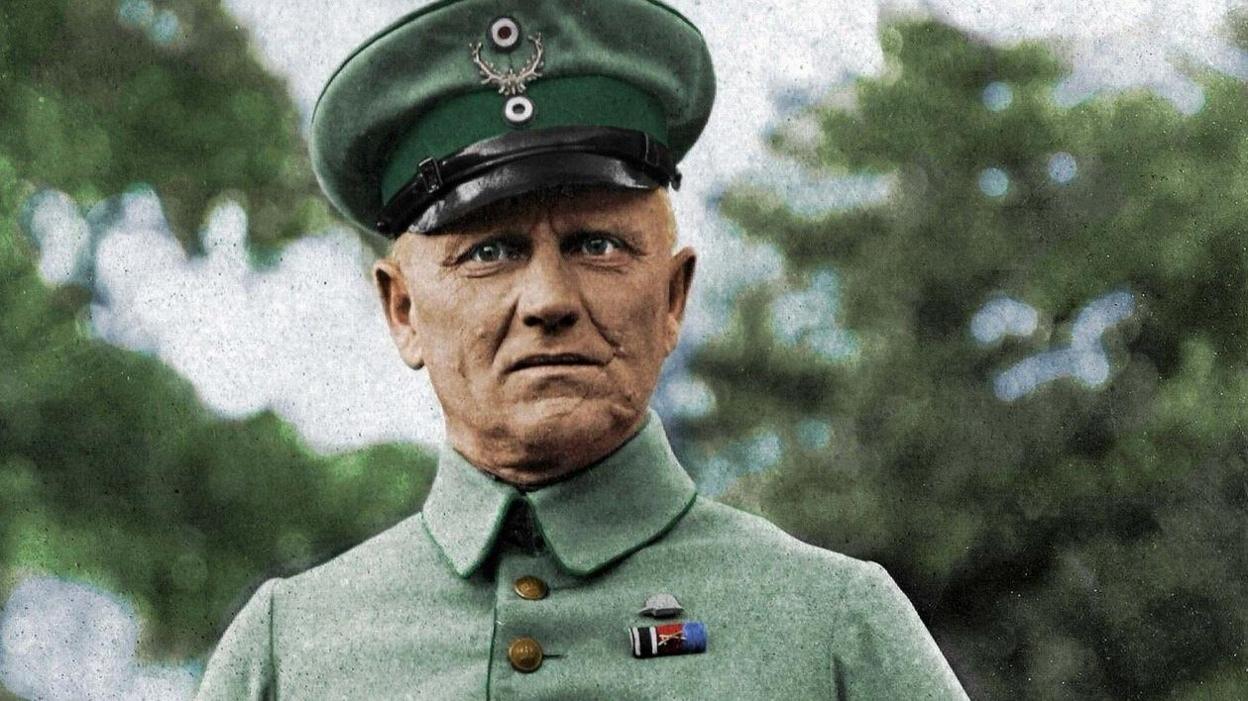 Lt Alexander Pfeifer, in a green German WW1 uniform with a peaked cap and a high collared jacket, stands in front of greenery