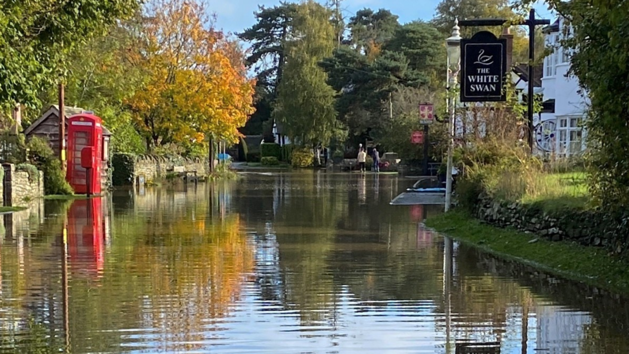 A flooded street with trees and a red phone box on the left-hand side and a white building with a black sign reading The White Swan on the right