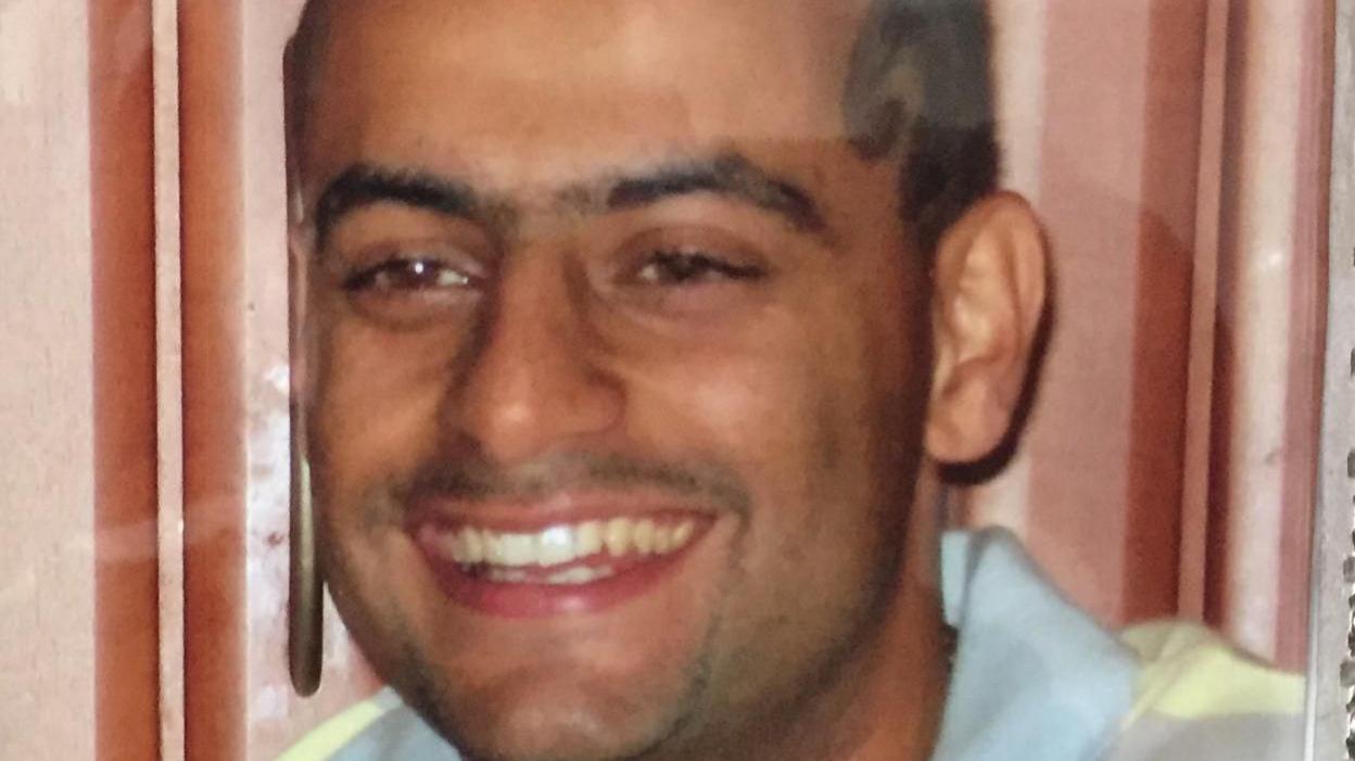 Pardeep Nagra smiling. He is in front of a brown wall and wearing a polo t-shirt. He has some styled facial hair and a pattern in his hair. 