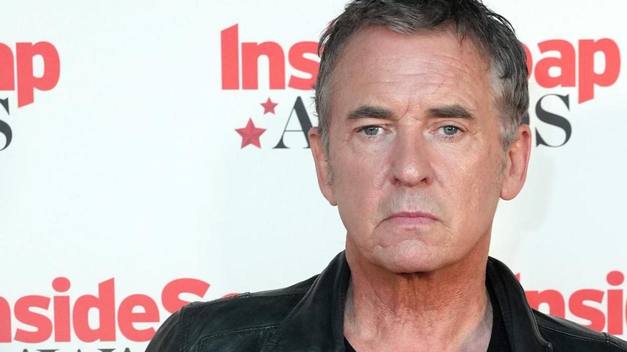 A man wearing a leather jacket and a black top standing in front of the press,
there is a backdrop behind him that reads inside soap awards 2024