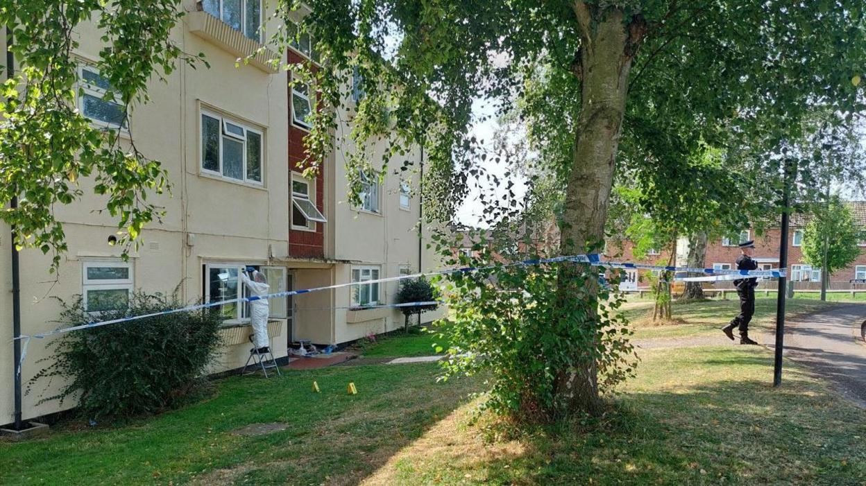 A sunny grass verge outside a block of flats cordoned off by blue and white police tape with a forensic officer and a police officer at the scene