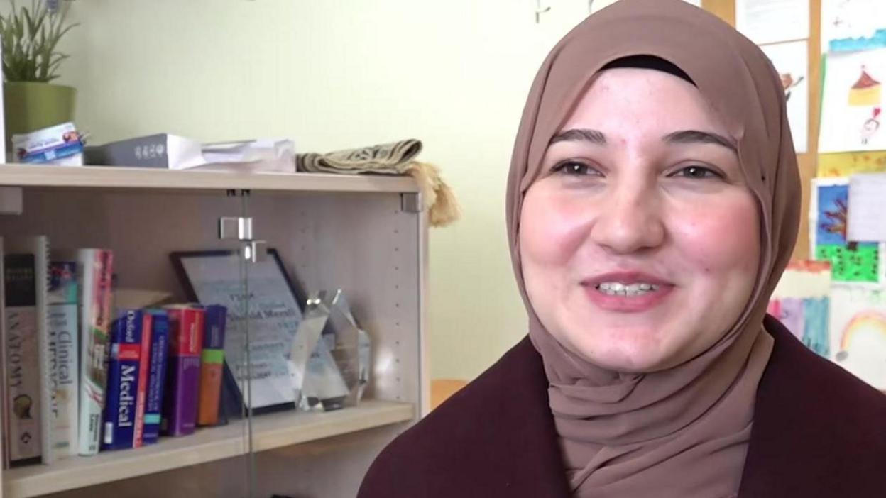 A woman in a pale brown head scarf and maroon top looks at the camera. She is sitting in an office. To her right is a glass-fronted cabinet, with books inside and a plant on top of it. The wall is pale yellow in colour.