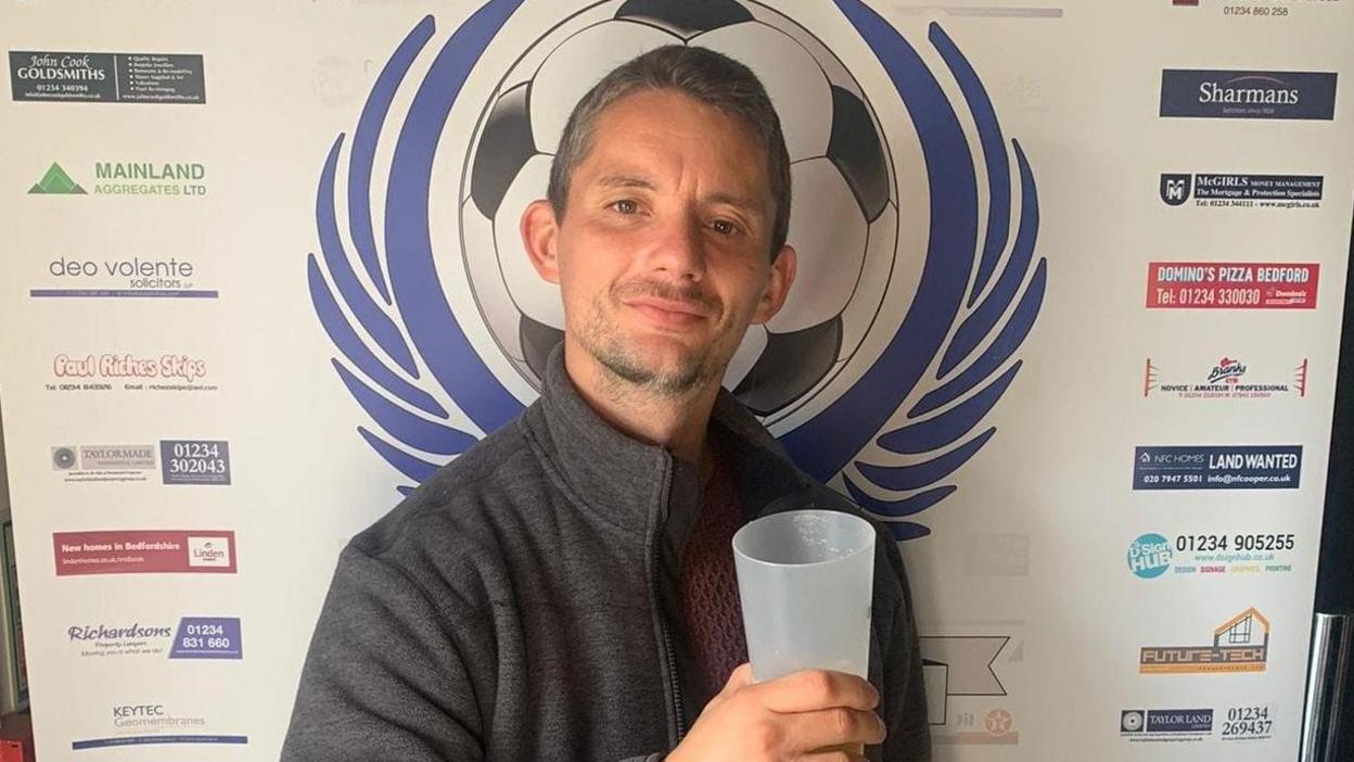 Christopher Allan stands in front of an advertising board. The board features a football surrounded by a blue laurel leaf. Mr Allan has short, grey hair and short beard and moustache. He is wearing a grey top and holding a plastic beaker.