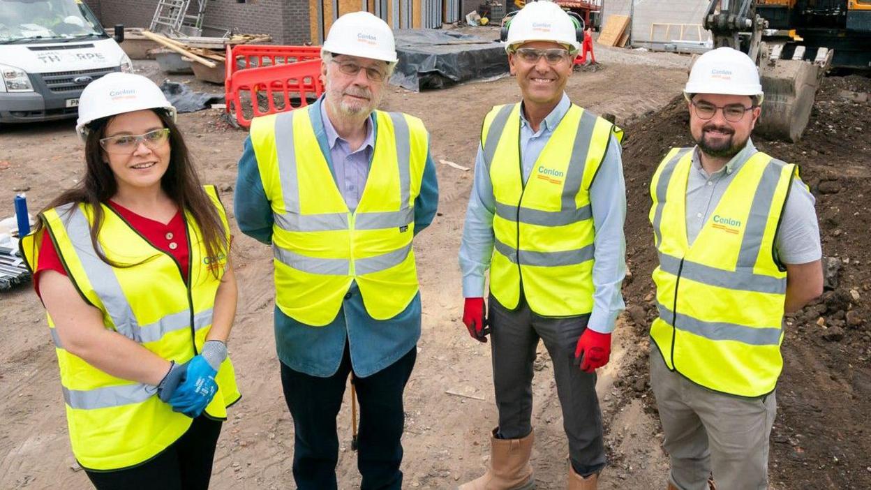 Three men and a woman stand on a building site wearing high-vis yellow vests, gloves and hard hats. They are all smiling. 