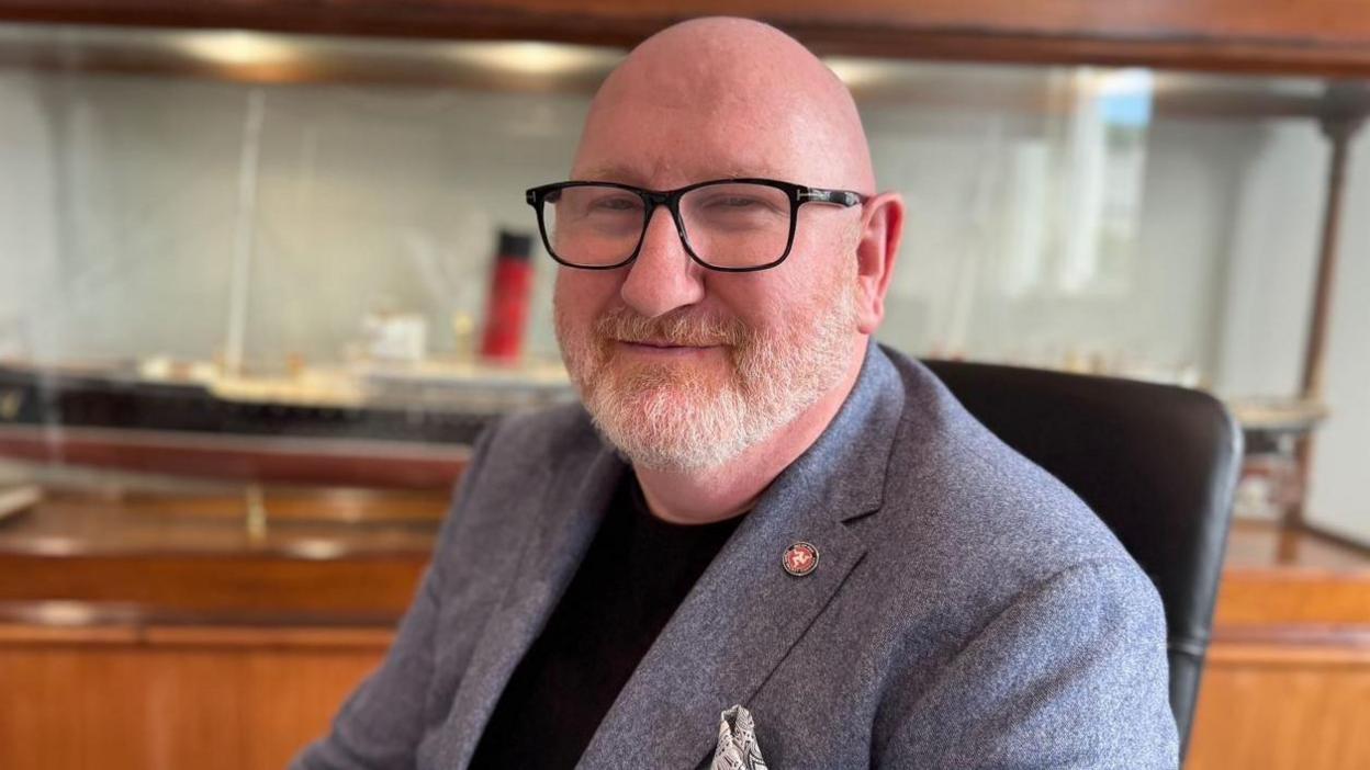 Brian Thomson is bald and has a grey short beard. He's wearing glasses and wearing a black top with a grey suit jacket.