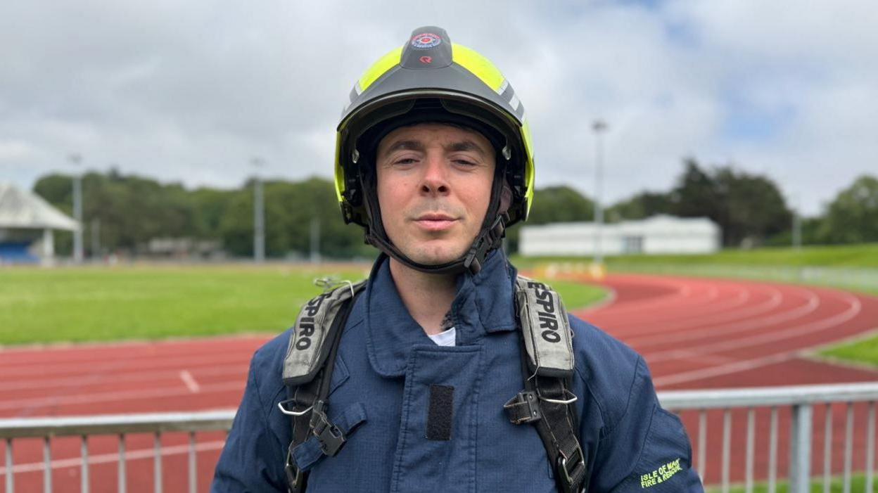 Nick Hays is wearing a blue fire fighter jacket, straps over his shoulders and yellow fluorescent helmet, standing in front of the orange tarmacked race track at the NSC.