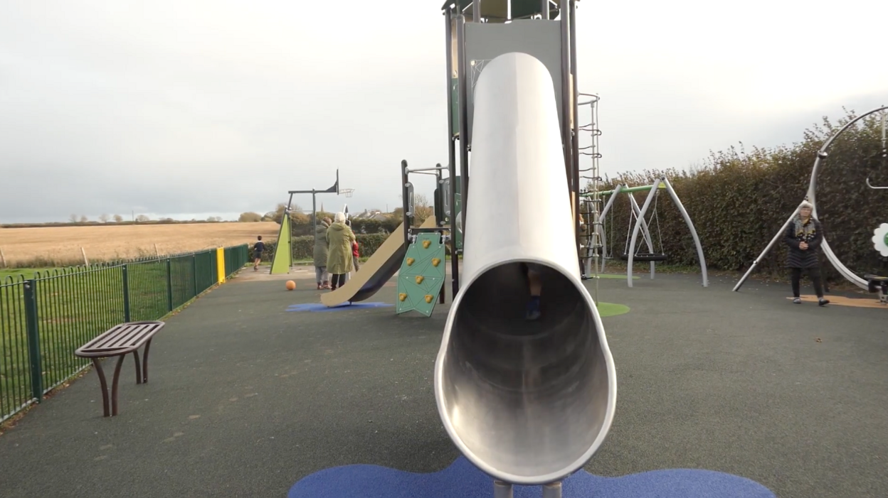 A new play area including a metal slide, rubber flooring and a range of apparatus. People can be seen playing and grey skies behind.