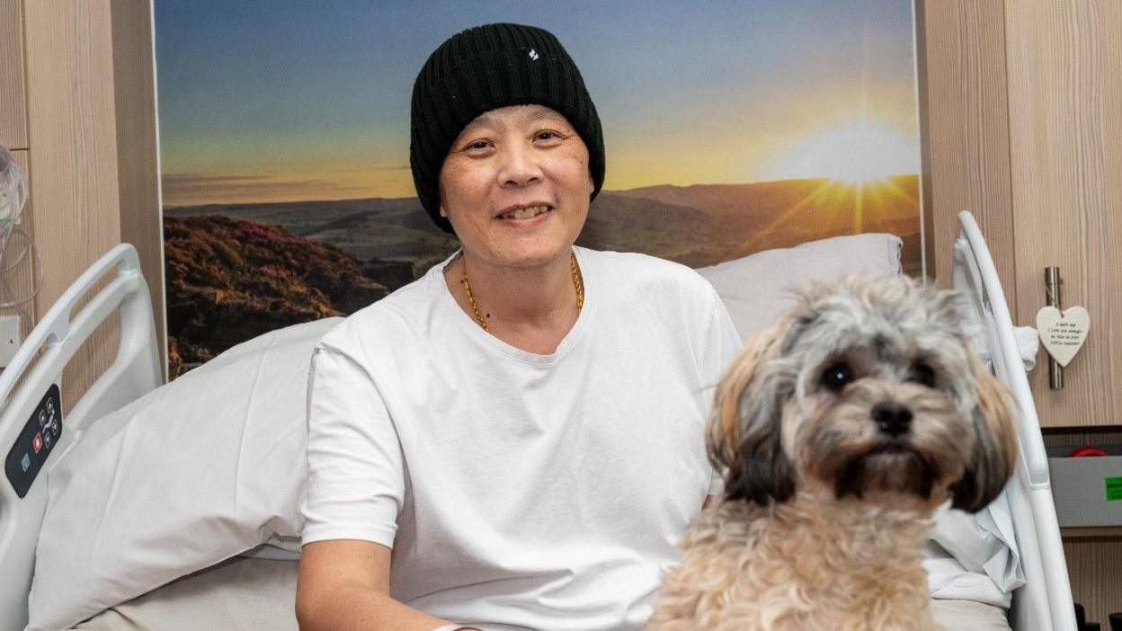 Lung Li with his pet dog both sat in a hospital-style bed with a poster of a sun lit landscape behind