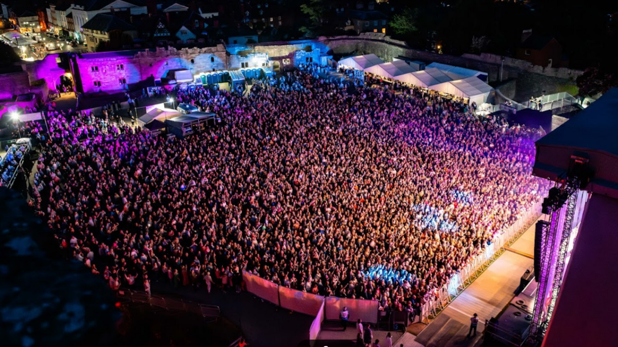 A shot taken from above the venue showing the whole audience bathed in purple and white light. To the right, modern stage rigging is visible, while around the other two sides of the auditorium, the castle walls and merchandise tents line the scene. In the distance, car and shop lights can be seen in the market square.