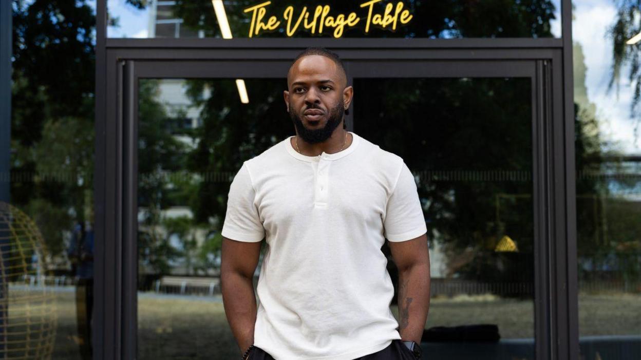 A young black man wearing a white T-shirt and standing outside in front of glass doors