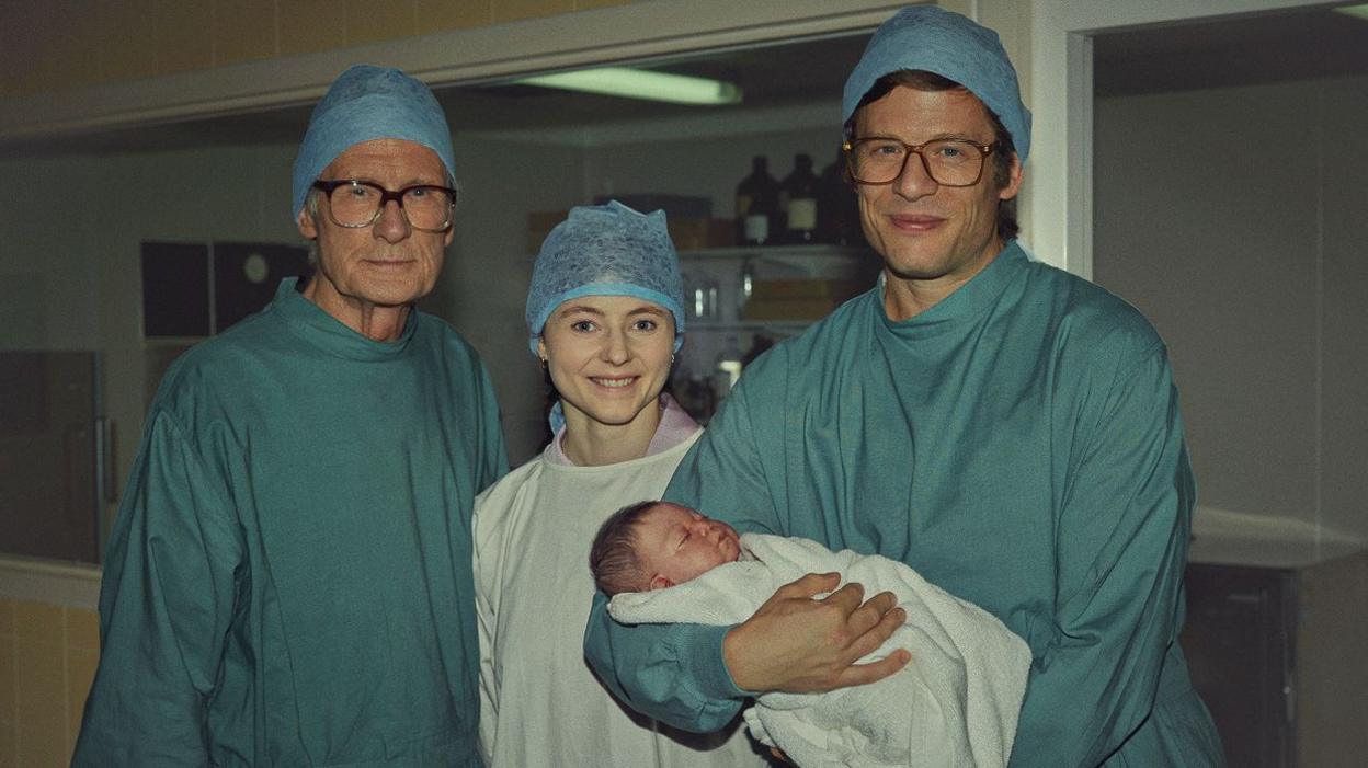 Bill Nighy, Thomasin McKenzie and James Norton are stood next to each other in blue scrubs. James is holding a baby wrapped in a white blanket.