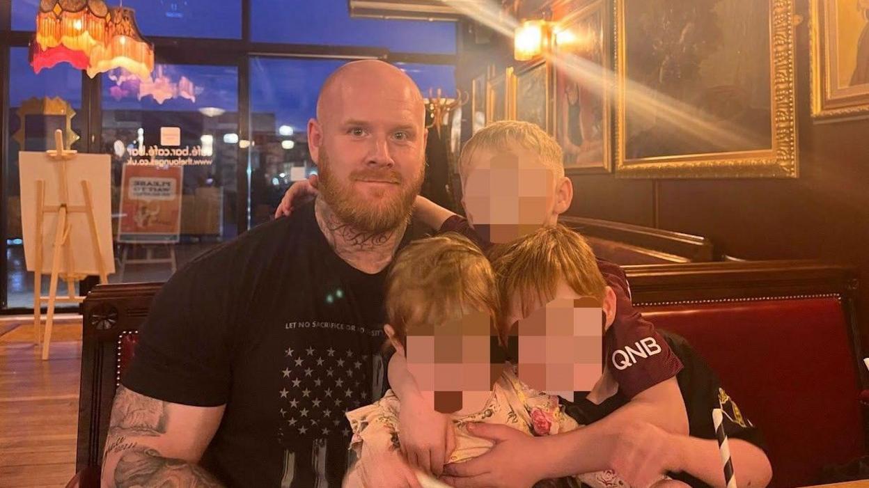 Lenny Scott, who has a shaved head and ginger beard, sits in a restaurant holding his three young children whose faces have been blurred