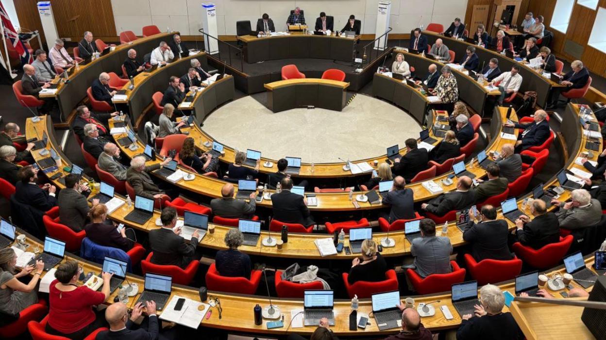 The Cornwall Council chamber with desks and seats arranged in a circle around the room. The photographer has taken the shot from the back of the room and we can see  microphones, laptops and paperwork on councillors' desks.