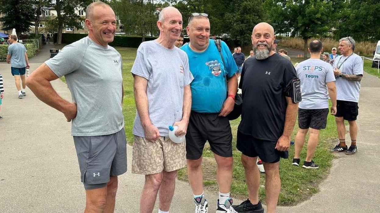 Four smiling men in shorts stood next to each other, looking past the camera at another photographer. Brendan is standing second from left wearing a grey T-shirt and shorts. There is grass and trees behind them.