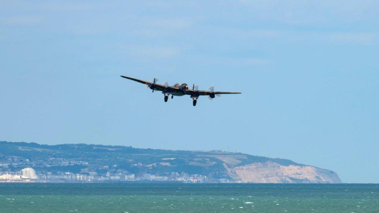 A plane flies low over a blue sea. Cliffs and a landmass are seen in the distance. 