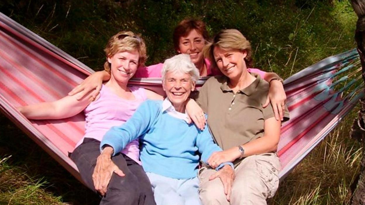 Sally Magnusson (left) in brown trousers and a pink T-shirt with sunglasses on her head, with her mother Mamie (centre), wearing a light blue jumper with white blouse underneath, sister Margaret (right) in beige trousers and a brown T-shirt. The three woman are sitting on a red and pink striped hammock with grass around them. Sally's sister Anna is crouching down behind them and is wearing a pink long-sleeved top.