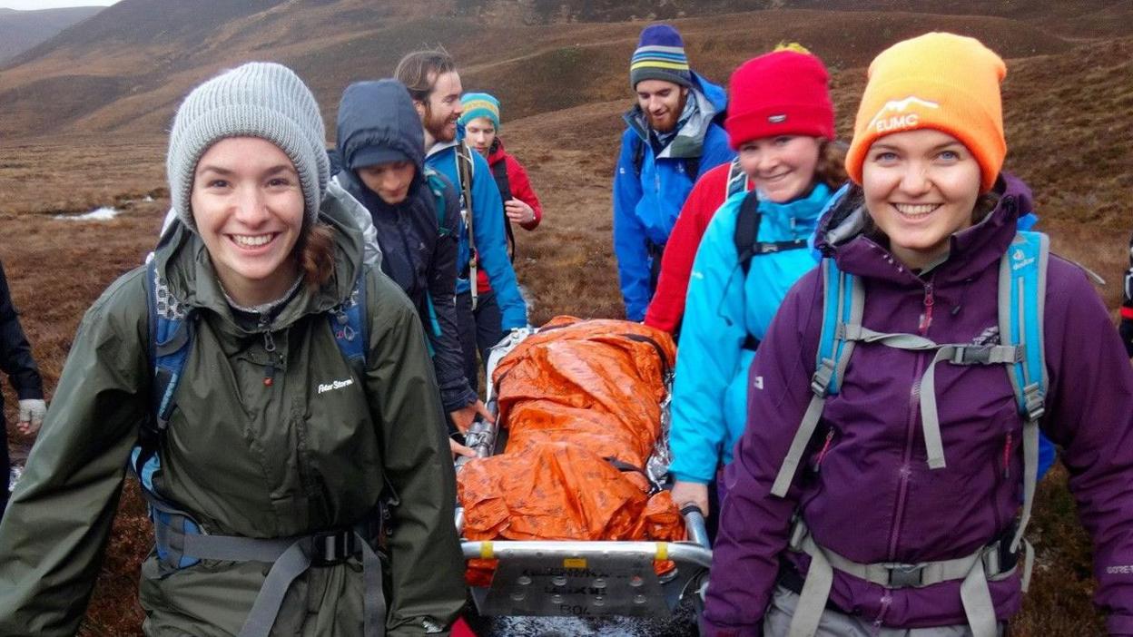 A group of young people wearing outdoor clothing carry a mountain rescue stretcher during a training exercise