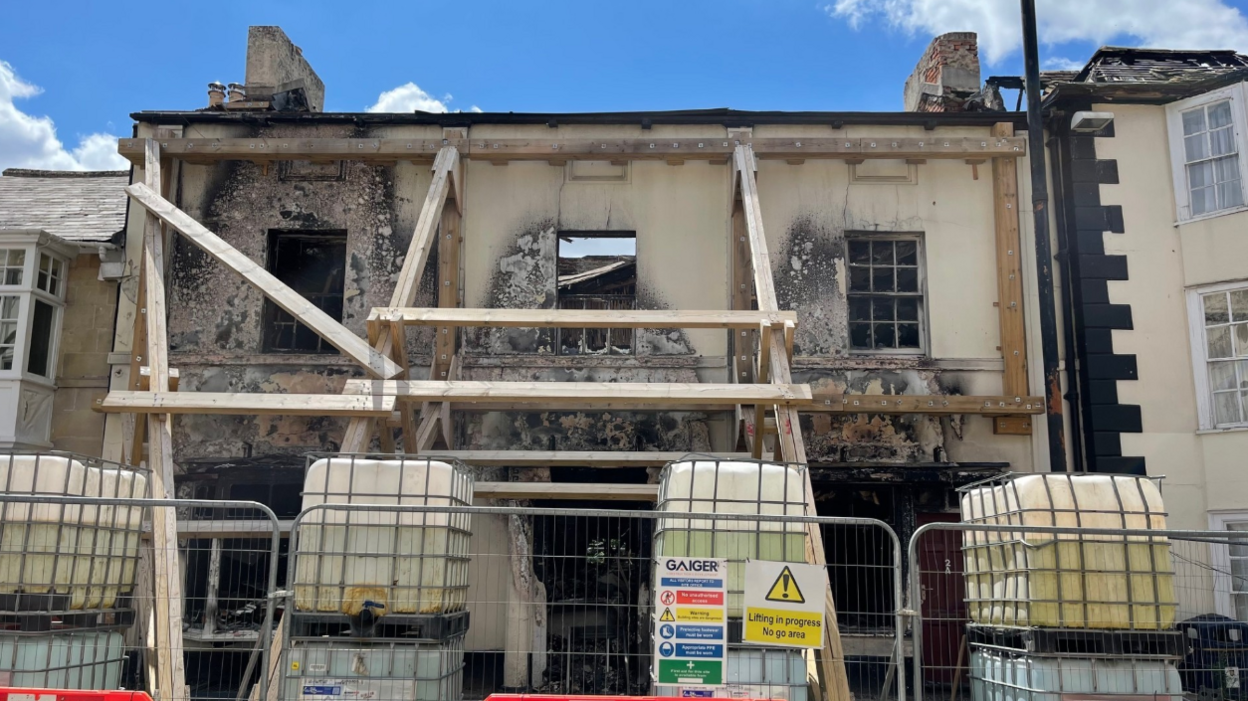 A building gutted by fire with blackened areas around several windows. Glass is missing from two upstairs windows. Four water tanks sit in front of the building, which is being supported by wooden scaffolding to prevent the facade from collapsing. A metal fence is apparent in the foreground.