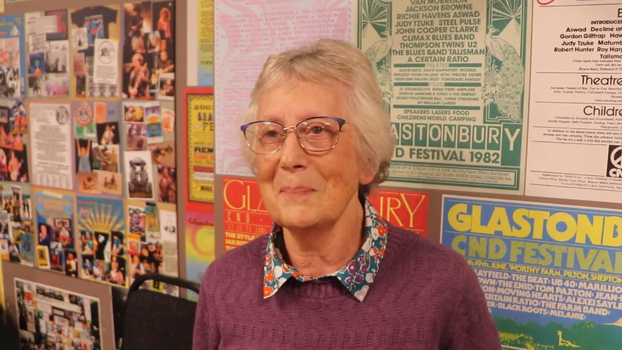An older woman is standing in front of a wall. She is wearing a purple jumper and glasses. On the wall are old Glastonbury posters and photos.