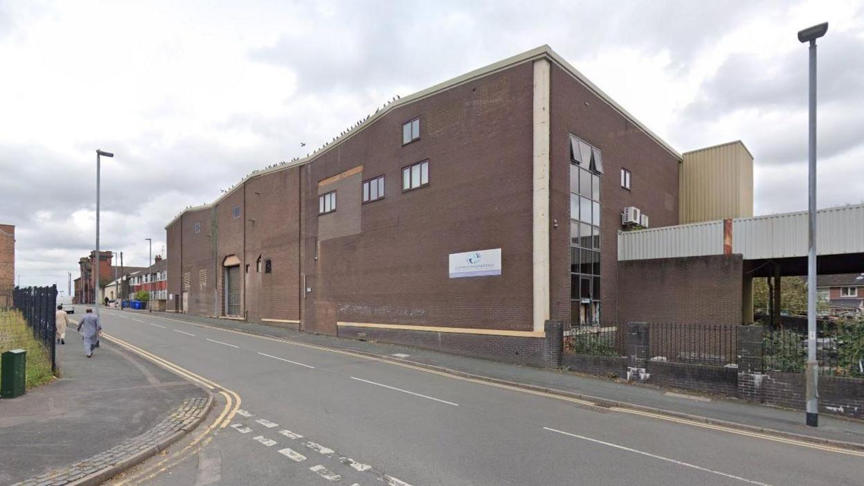 A Google street view image of an old disused brown factory building at the side of a road. People can be seen walking along the pavement on the right.