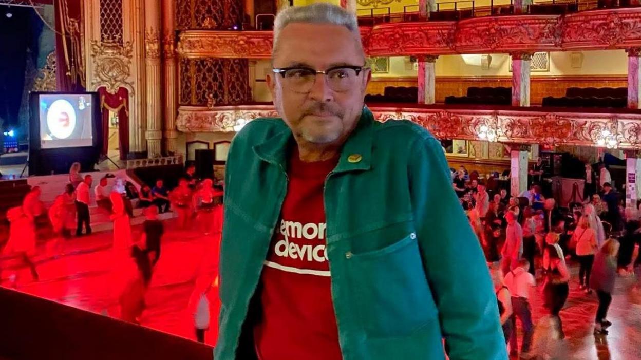 a man with grey hair and glasses wearing a green jacket and red top.