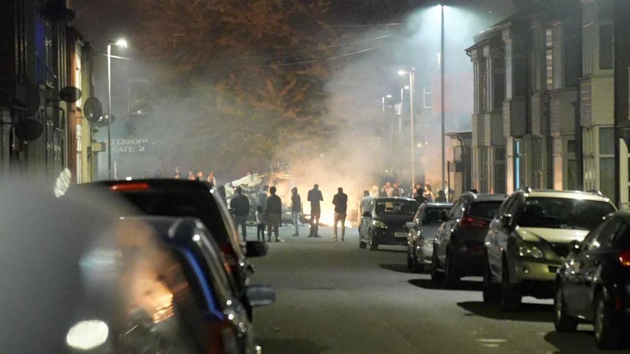 A residential street at night, with cars parked on both sides of the road. In the distance, a group of people stand around the remains of a burning bus.