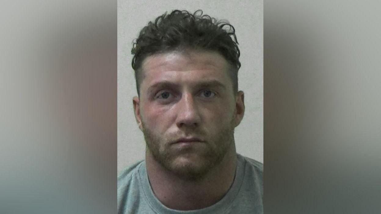 Jake McIntyre mugshot. He has short dark curly hair, a neat stubble beard and is wearing a grey sweatshirt.