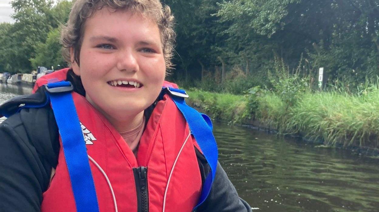 A teenage girl with short brown hair, canoeing on a river and wearing a red life vest. She is smiling.