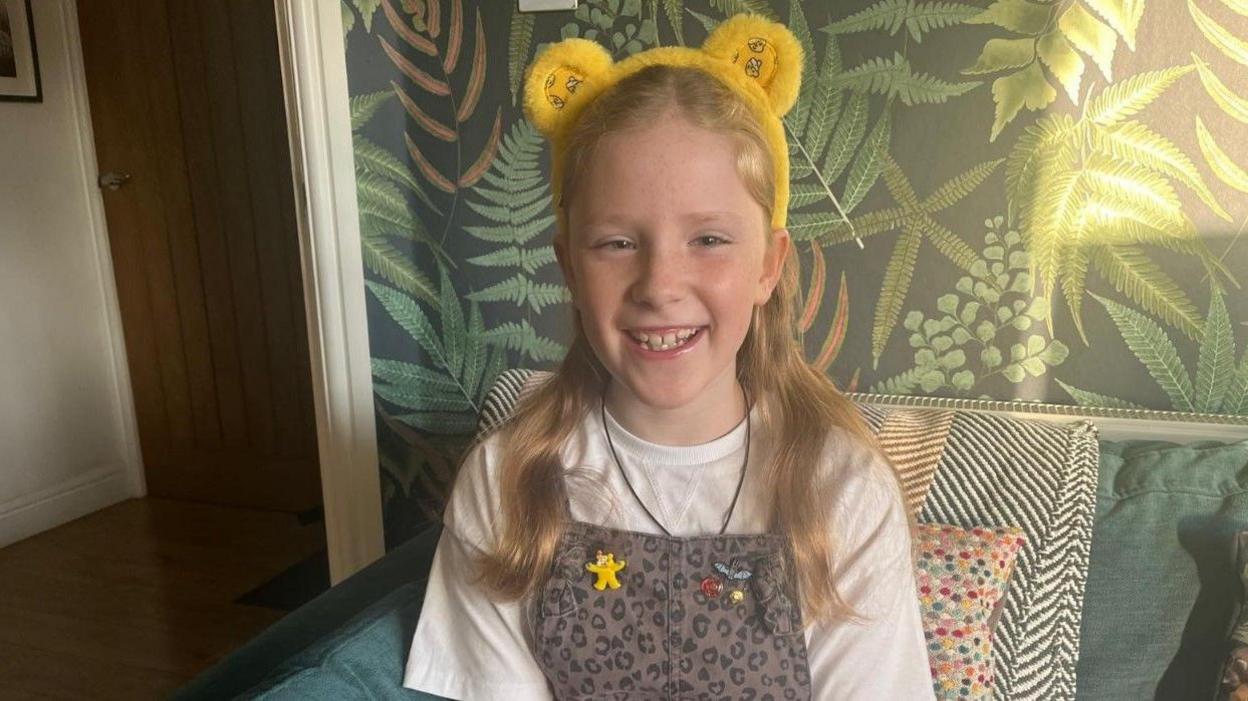 Hannah, smiling with long hair, wearing Children in Need mascot Pudsey Bear ears. She is sitting down, with a door and a green-coloured wall behind her.